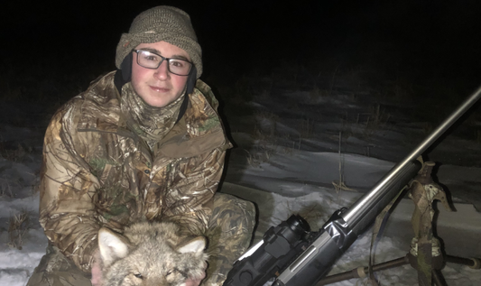 A young man is sitting on the snowy ground, holding a dead coyote