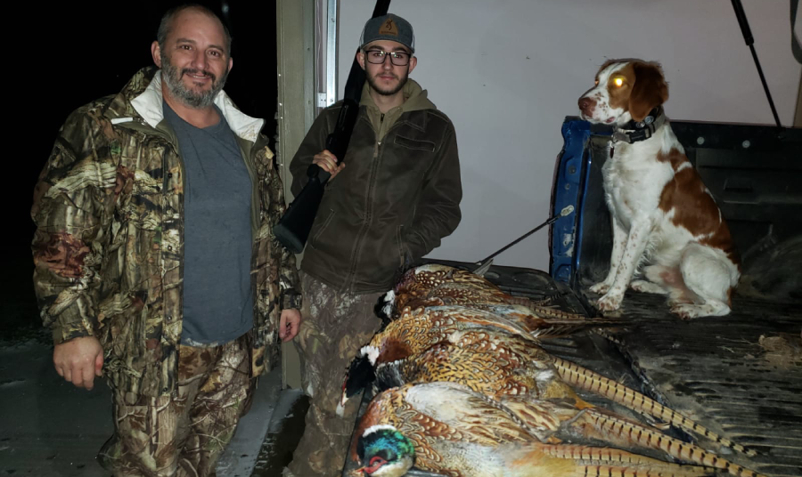 two hunters posing with their hunting dog next to a pickup truck, which has a collection of pheasants laid out on its bed.